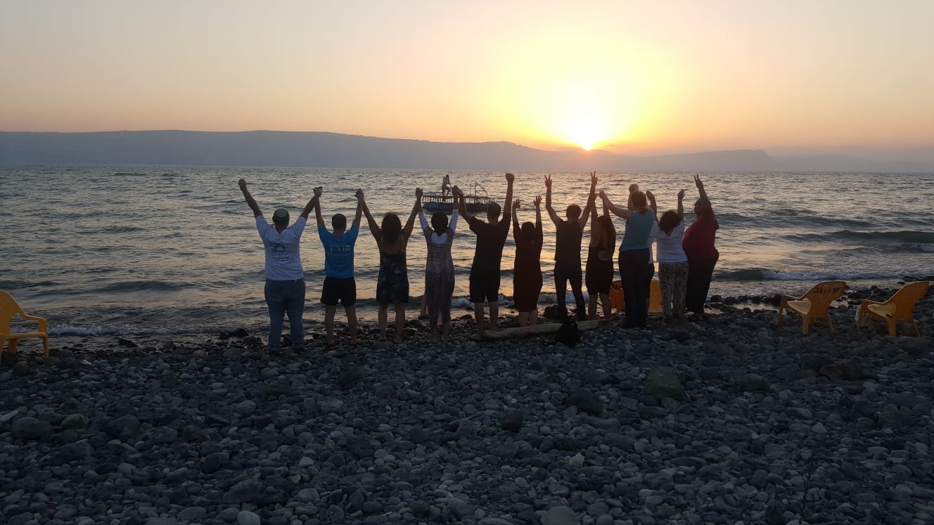 12 אנשים עומדים על שפת הכנרת עם הגב למצלמה ומרימים ידים לשמים קלאבהאוס טבריה